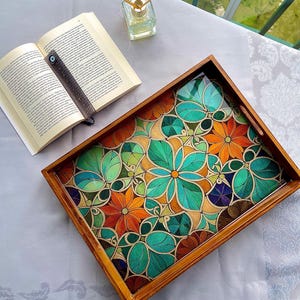 May include: A wooden serving tray with a vibrant floral pattern in orange, green, and blue. The tray has a clear, glossy surface and a dark brown wooden frame. An open book and a small glass bottle with reeds are also visible.