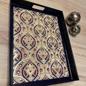 May include: Rectangular serving tray with a navy blue frame and handles. The tray's interior features a cream-colored background with a repeating pattern of red, blue, and silver floral designs. Two silver decorative spheres are next to the tray.