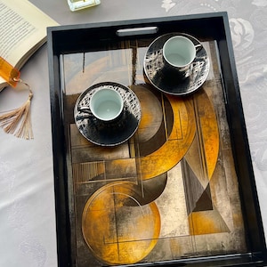 May include: A black rectangular serving tray with a geometric design in gold, brown, and gray. Two black teacups and saucers with a silver pattern are on the tray. The tray has handles and a glossy finish.