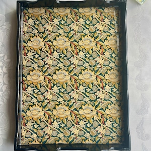 Könnte beinhalten: Rechteckiges Serviertablett mit glänzendem schwarzem Rahmen und Griffen. Das Innere des Tabletts weist ein Blumenmuster in Grün-, Gelb- und Rottönen auf. Das Tablett ist zum Servieren von Speisen oder Getränken bestimmt.