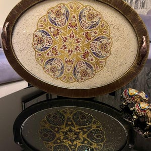 Könnte beinhalten: Ein ovales dekoratives Tablett mit goldfarbenem Rand und Griffen. Das Innere des Tabletts weist eine cremefarbene Oberfläche mit einem detaillierten floralen Design in Gold, Rot und Blau auf. Das Tablett wird auf einer schwarzen Oberfläche mit einer Spiegelung des Tabletts darunter präsentiert.