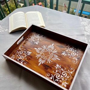 May include: A rectangular wooden serving tray with a dark brown stained interior and white painted edges. The tray features a white floral design. An open book rests on a white tablecloth next to the tray.