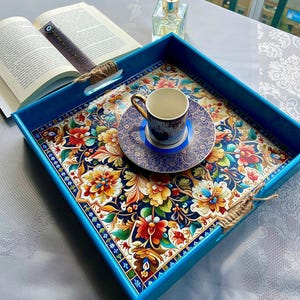 May include: A blue serving tray with a floral pattern in red, yellow, and blue. A small teacup and saucer are on the tray. An open book and a diffuser are in the background.