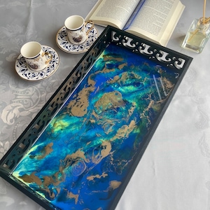 May include: A rectangular serving tray with a black frame and a vibrant, abstract design in shades of blue, gold, and green. Two small coffee cups and saucers with blue and white patterns are on the left. An open book and a diffuser are also present.