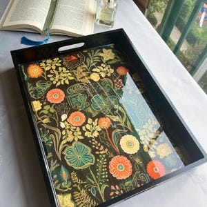 Pode incluir: Uma bandeja de servir retangular preta com um design floral. A bandeja apresenta um fundo escuro com flores laranja, amarelas e verdes. Um livro aberto com um marcador azul e um difusor de vidro também são visíveis.
