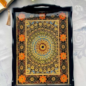 May include: Black serving tray with a colorful, intricate design. The tray features a central mandala pattern in gold, blue, and yellow, surrounded by orange starburst motifs. The tray has a glossy finish and integrated handles.