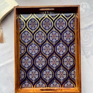 May include: Wooden serving tray with a rectangular shape. The tray features a decorative pattern in gold, blue, and white. The tray has a wooden frame with cut-out handles on the sides.