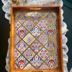 Puede incluir: Bandeja de madera para servir con un diseño de azulejos coloridos. La bandeja presenta un marco de madera marrón claro y detalles dorados. El diseño interior incluye patrones florales rojos, azules y amarillos. Se ven dos pequeñas figuras decorativas de elefantes.