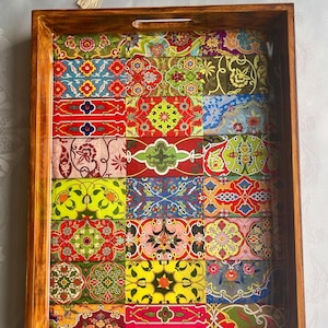 May include: Wooden serving tray with a mosaic design. The tray has a dark brown wooden frame and a colorful interior with a patchwork of floral and geometric patterns in red, green, yellow, and blue. The tray has cut-out handles.