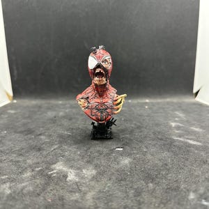 May include: A detailed bust of a zombie Spider-Man, featuring a red and black costume with exposed flesh and bone. The figure is mounted on a black base, showcasing intricate details and a distressed appearance. The bust is a collectible item.