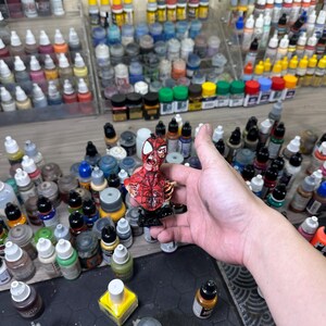 May include: A detailed, hand-painted miniature of Spider-Man in a dynamic pose. The figure is predominantly red with black web detailing. The background features a collection of various paint bottles, suggesting a hobby or craft setting.