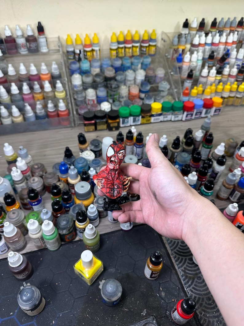 May include: A detailed, hand-painted miniature of Spider-Man in a dynamic pose. The figure is predominantly red with black web detailing. The background features a collection of various paint bottles, suggesting a hobby or craft setting.