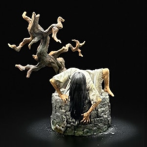 May include: A detailed figurine depicting a horror scene. A figure with long black hair emerges from a stone well, with a gnarled tree above. The figurine is painted in muted tones, with the X-Art Studio logo in the top right corner.