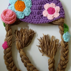 Pode incluir: Chapéu de crochê roxo com uma cauda trançada de fio marrom e enfeites de flores coloridas. O chapéu tem uma flor rosa, azul e laranja no topo e uma flor rosa no lado. A cauda trançada tem flores verdes, azuis e laranja.
