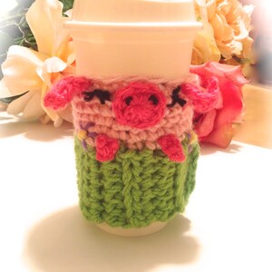 May include: A crocheted coffee cup cozy featuring a pink pig face with black eyes and a pink nose. The cozy is green with pink accents and a pink flower design.