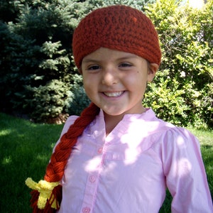 May include: A young person wearing a pink button-down shirt with a long, braided, red and yellow yarn hairpiece. The hairpiece is styled with a yellow flower at the end. The person is also wearing a crocheted brown hat.