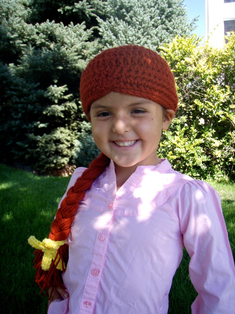 May include: A young person wearing a pink button-down shirt with a long, braided, red and yellow yarn hairpiece. The hairpiece is styled with a yellow flower at the end. The person is also wearing a crocheted brown hat.
