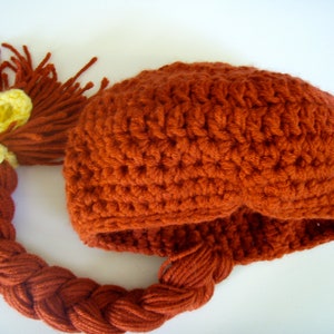 May include: A crocheted hat with a long, braided tail. The hat is a reddish-brown colour and the tail is braided with a yellow ribbon at the top.