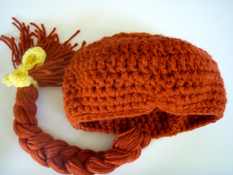 May include: A crocheted hat with a long, braided tail. The hat is a reddish-brown colour and the tail is braided with a yellow ribbon at the top.