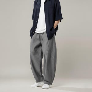 May include: A casual outfit featuring a navy blue short-sleeved button-down shirt over a white t-shirt, paired with gray, loose-fitting pants. The pants have a drawstring waist and are worn with white sneakers. The outfit is simple and comfortable.
