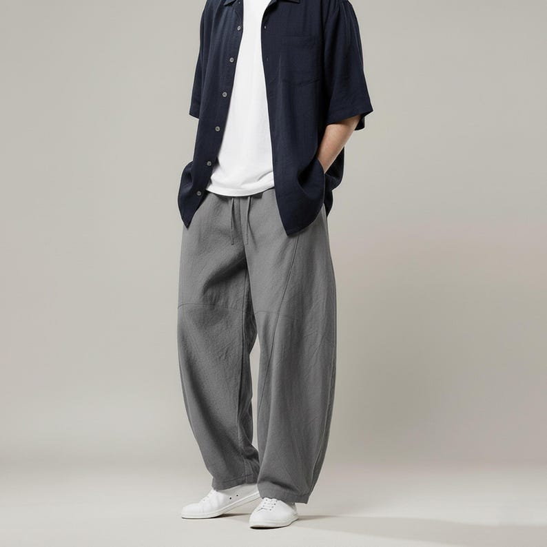 May include: A casual outfit featuring a navy blue short-sleeved button-down shirt over a white t-shirt, paired with gray, loose-fitting pants. The pants have a drawstring waist and are worn with white sneakers. The outfit is simple and comfortable.