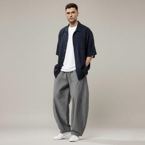 May include: A person wearing a navy blue short-sleeved button-down shirt over a white t-shirt, paired with gray, loose-fitting pants and white sneakers. The outfit is casual and modern, suitable for everyday wear.