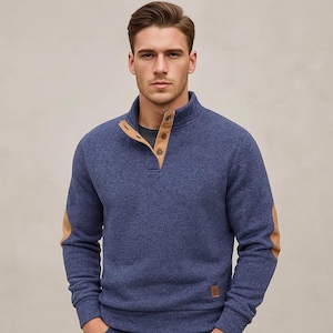 May include: A blue pullover sweater with a high collar and three buttons. The sweater has tan elbow patches and a small tan patch near the bottom. The model is wearing a gray t-shirt underneath.