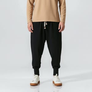 May include: Black casual pants with a drawstring waist and ribbed cuffs. The pants are paired with a long-sleeved tan shirt and white sneakers with brown soles. The outfit is simple and comfortable.