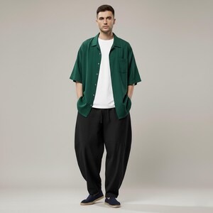 May include: A person wearing a green short-sleeve button-down shirt over a white t-shirt, paired with black, loose-fitting pants. The outfit is completed with navy blue slip-on shoes. The background is a solid, neutral gray.
