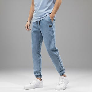 May include: Light blue denim joggers with a drawstring waist and elastic cuffs. The pants feature a small square patch on the front pocket. The model is wearing white sneakers and a light blue t-shirt.