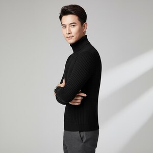 May include: A black ribbed turtleneck sweater. The sweater has a high neckline and long sleeves. The person is wearing gray pants and a watch. The background is a solid light gray.