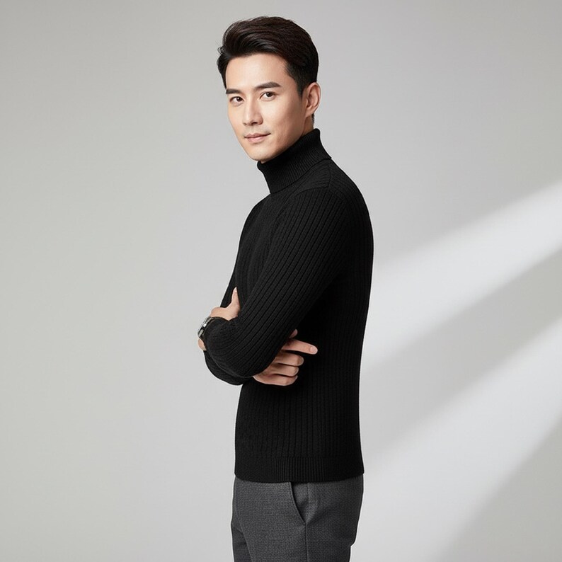 May include: A black ribbed turtleneck sweater. The sweater has a high neckline and long sleeves. The person is wearing gray pants and a watch. The background is a solid light gray.