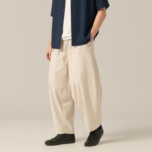 May include: Beige, wide-leg pants with a relaxed fit. The pants have a drawstring waist and are paired with a dark blue short-sleeve shirt and black sneakers. The outfit is casual and comfortable.