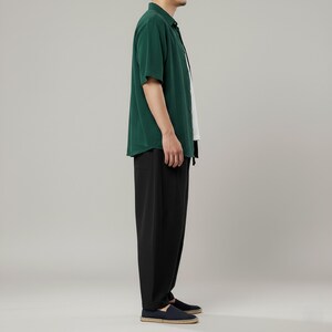 May include: A person wearing a dark green short-sleeved button-down shirt, a white undershirt, and black pants. The person is also wearing dark blue shoes. The outfit is casual and simple.