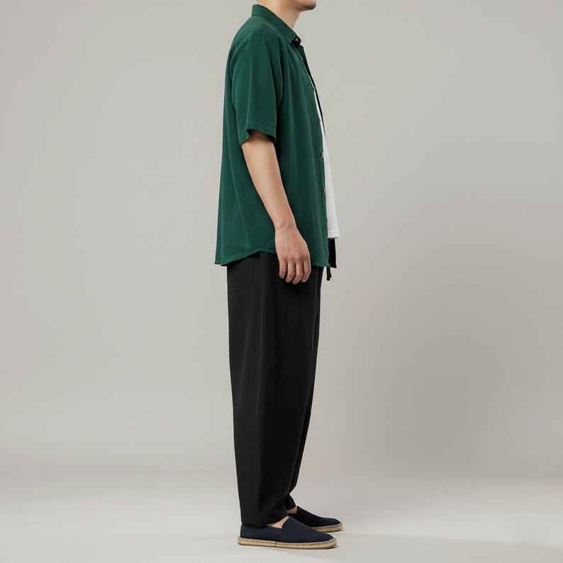 May include: A person wearing a dark green short-sleeved button-down shirt, a white undershirt, and black pants. The person is also wearing dark blue shoes. The outfit is casual and simple.