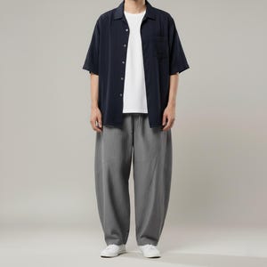 May include: A person wearing a navy blue short-sleeved button-down shirt over a white t-shirt, paired with gray, loose-fitting pants and white sneakers. The outfit is casual and modern.