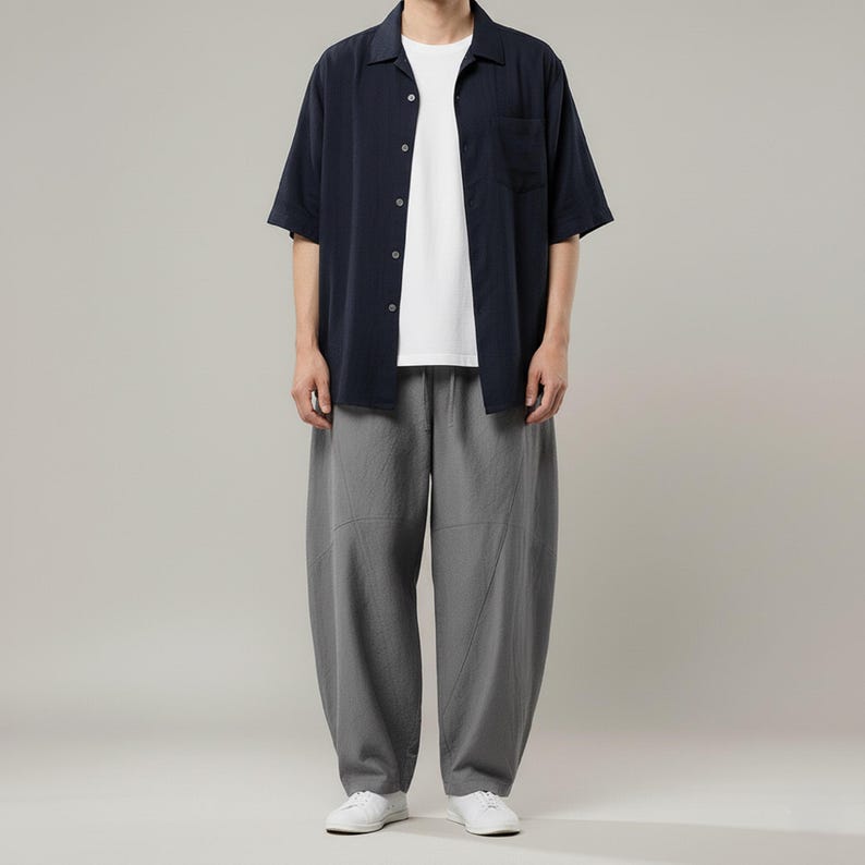 May include: A person wearing a navy blue short-sleeved button-down shirt over a white t-shirt, paired with gray, loose-fitting pants and white sneakers. The outfit is casual and modern.