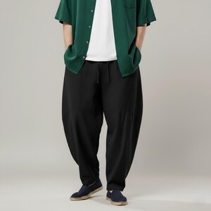 May include: Black, loose-fitting pants with a drawstring waist. The pants are paired with a green button-down shirt and dark blue shoes. The style is casual and comfortable.