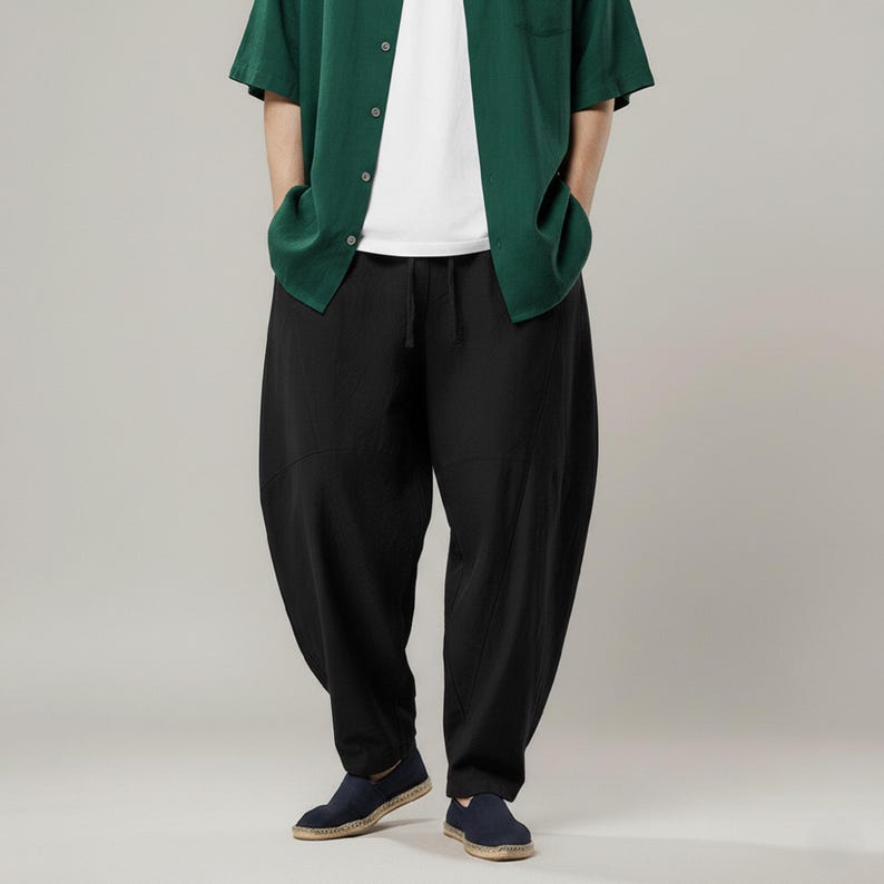 May include: Black, loose-fitting pants with a drawstring waist. The pants are paired with a green button-down shirt and dark blue shoes. The style is casual and comfortable.