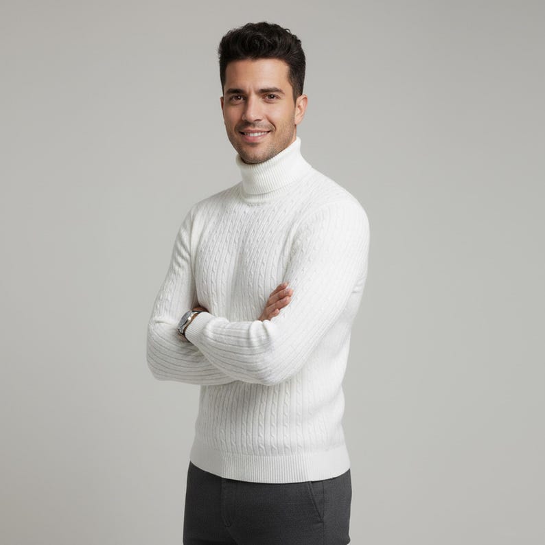May include: A white cable-knit turtleneck sweater. The sweater has a ribbed hem and cuffs. The person is wearing a silver watch and gray pants. The background is a solid light gray.