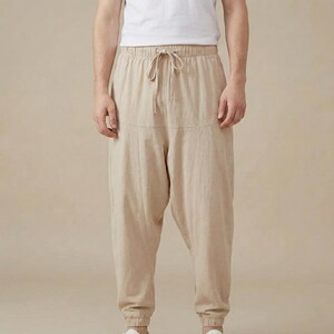 Puede incluir: Pantalones de lino beige con cintura con cordón y puños elásticos. Los pantalones tienen un corte holgado y un sutil detalle de costura en la parte delantera. El modelo lleva una camiseta blanca. Adecuado para uso casual.