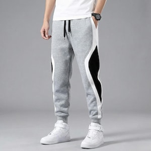 Op de afbeelding: Grijze joggingbroek met witte en zwarte zijpanelen. De broek heeft een tailleband met trekkoord en geribbelde enkels. De persoon draagt witte sneakers en een wit t-shirt. De joggingbroek heeft een casual stijl.