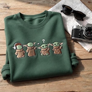 May include: Green sweatshirt featuring an embroidered design of four Grogu characters in various poses. The characters are wearing brown robes and have large ears. One character has headphones. A question mark is above one character's head.