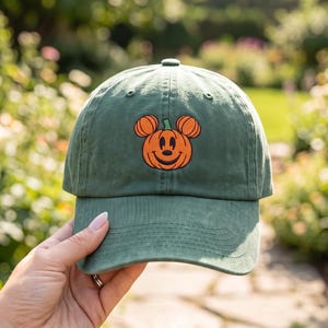 Peut inclure: Une casquette de baseball vert sauge avec une broderie orange représentant une citrouille en forme de tête de souris. La casquette présente une visière courbée, une calotte structurée avec un bouton supérieur et des œillets de ventilation.