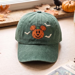 May include: A forest green baseball cap featuring an embroidered orange pumpkin shaped like a mouse head with a smiling face. The design includes small white embroidered bats surrounding the pumpkin. The cap has a curved brim and stitched eyelets.