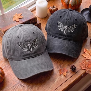 May include: Two charcoal gray baseball caps featuring an embroidered moth design with a skull pattern on the thorax. Each cap has a curved brim, a button on the crown, and stitched eyelets. The caps are made of a washed, distressed fabric.