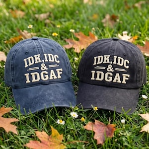 May include: Two baseball caps, one in navy blue and one in charcoal gray, each featuring the embroidered white text IDK, IDC & IDGAF in a bold, block-style font. The caps have a structured crown, a curved brim, and a classic six-panel design.