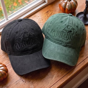 May include: A black baseball cap and a green baseball cap, both featuring embroidered text that reads Witchling, accompanied by small moon, star, and bat icons. The caps have a structured crown, a curved brim, and visible stitching details.