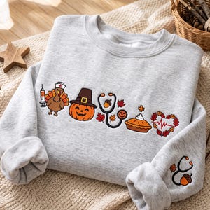 May include: A light grey crewneck sweatshirt featuring a graphic print of a turkey in a nurse cap with a syringe, a jack-o'-lantern, a stethoscope with an acorn, a pumpkin pie, and a heart with an EKG line. A stethoscope and acorn are on the sleeve.