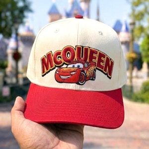 May include: A cream-colored baseball cap with a red brim featuring an embroidered graphic of a red race car and the text MCQUEEN in bold red letters with a dark outline. The cap has a structured crown and a button on the top center.